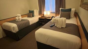 Quarto de hotel com duas camas, mesa de cabeceira com luminárias, janela com cortinas e espelho na parede.