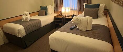 Ein Hotelzimmer mit zwei Betten, einem Nachttisch mit Lampen, einem Fenster mit Vorhängen und einem Spiegel an der Wand.