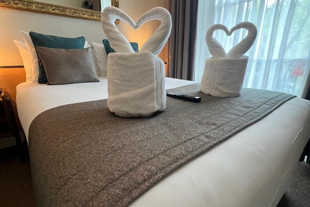 A hotel room with a neatly made bed, a heart-shaped towel arrangement, and a large mirror above the bed.