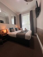 A hotel room with a large bed, a bedside table with a lamp, a flat-screen TV mounted on the wall, and a window with curtains.