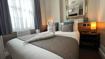 Ein Hotelzimmer mit einem ordentlich bezogenen Bett, einem großen Spiegel, einer Nachttischlampe und Blick ins Freie durch das Fenster.