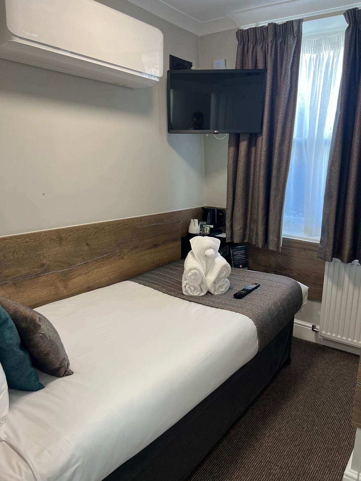 A hotel room with a single bed, a flat-screen TV mounted above it, a small table with a coffee maker, and a window with curtains.