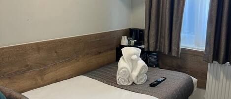 A hotel room with a single bed, a flat-screen TV mounted above it, a small table with a coffee maker, and a window with curtains.