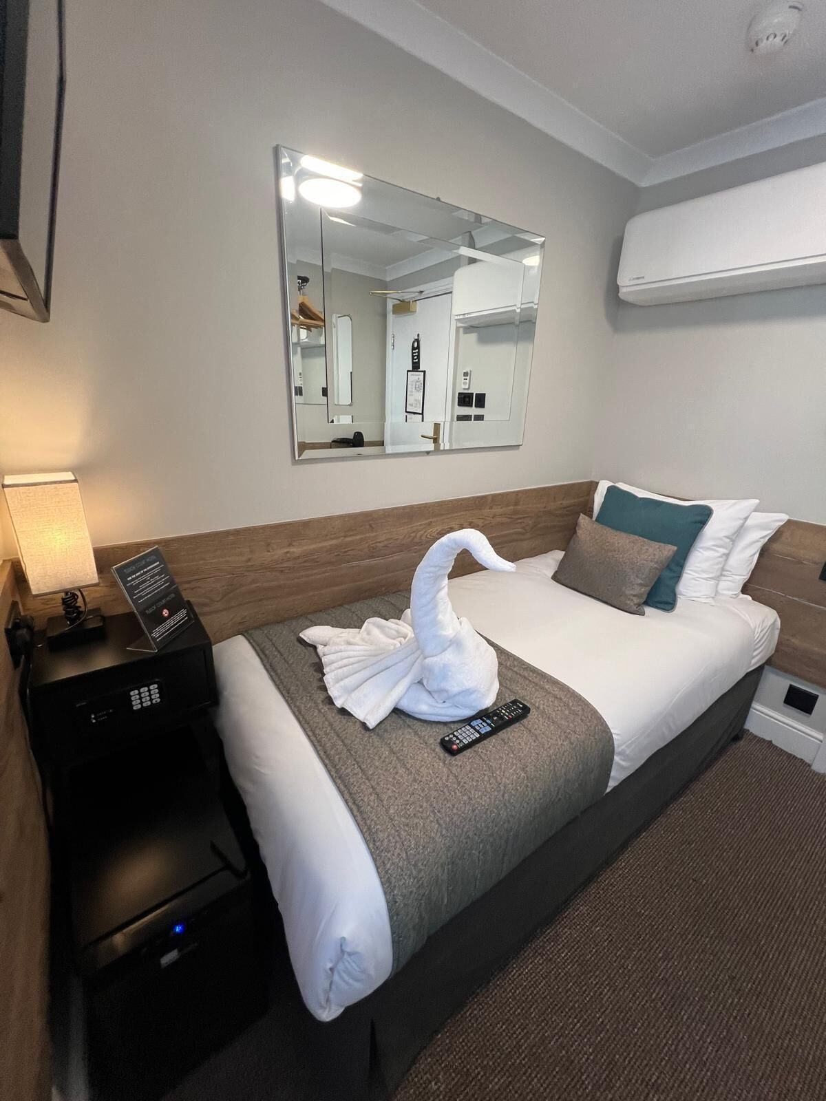 A hotel room with a bed, a nightstand, a remote control, a towel shaped like a swan, and a mirror on the wall.