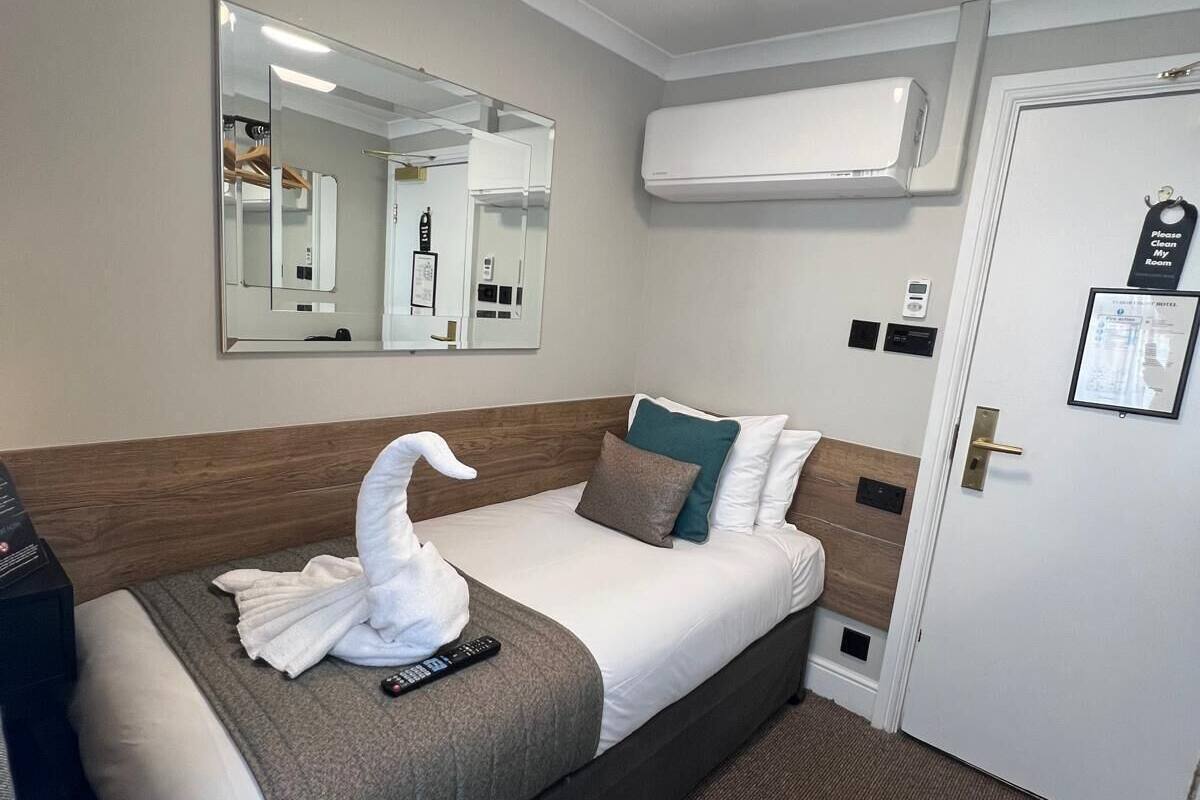 A hotel room with a bed, a mirror, an air conditioning unit, a door with a sign, and a remote control on the bed.