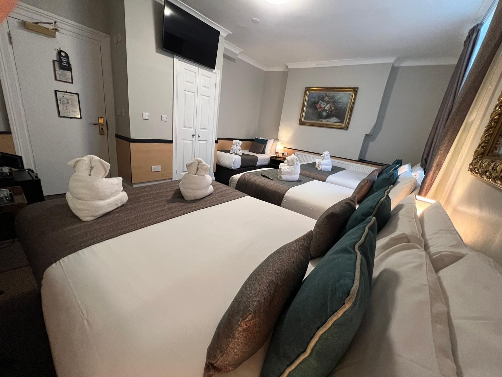 Een hotelkamer met twee bedden, een flatscreen televisie boven het linkerbed, een schilderij aan de muur en een deur met naambord.