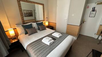 A hotel room with a large bed, a mirror, a wardrobe, and a desk with a lamp.