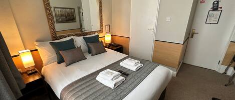 A hotel room with a large bed, a mirror, a wardrobe, and a desk with a lamp.
