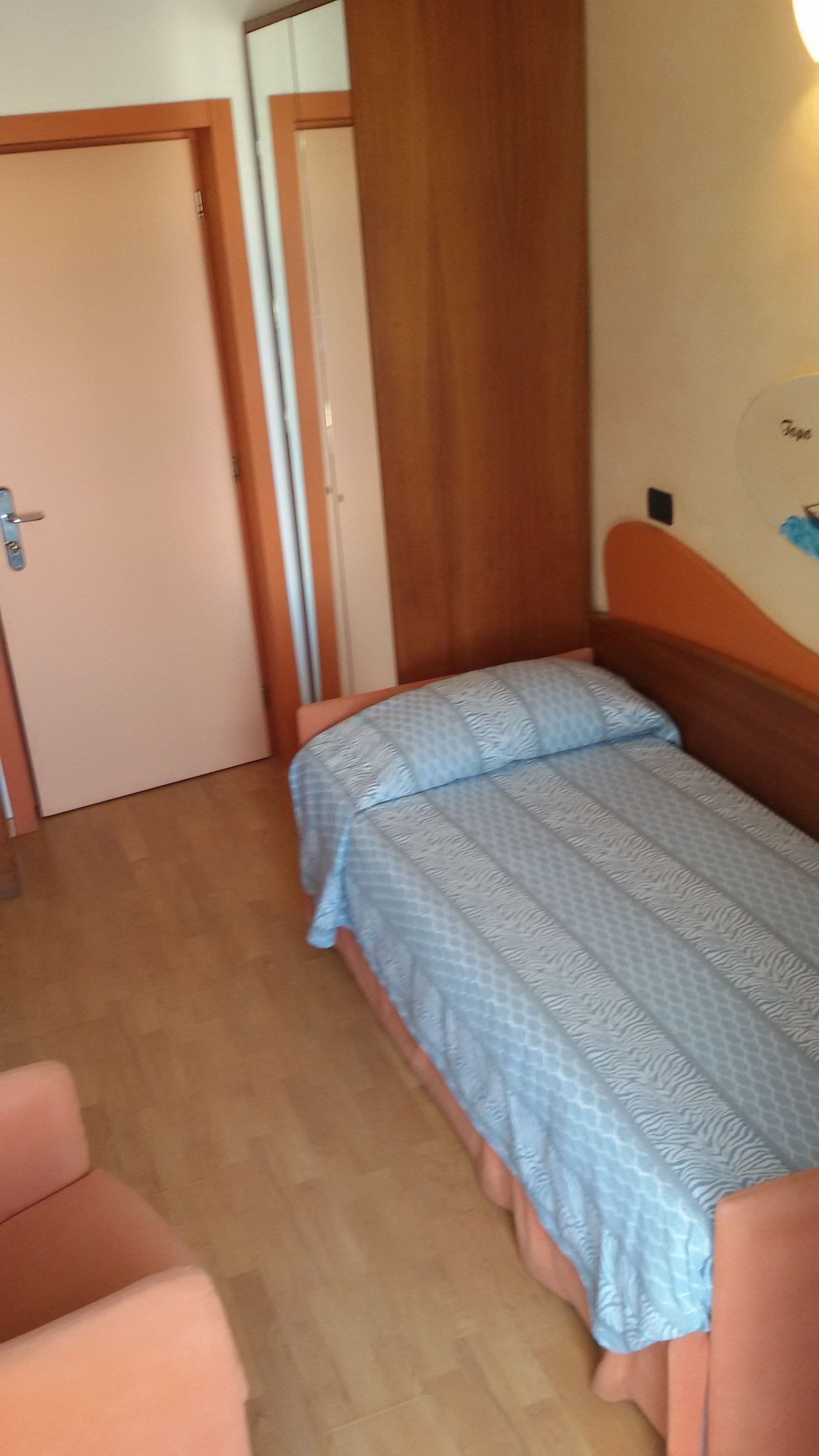 Een kleine kamer met een enkel bed, een houten hoofdeinde, een deur en een wandlamp.