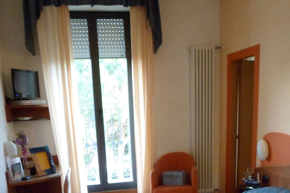 A bedroom with a bed, a desk with a chair, a radiator, and a window with curtains.