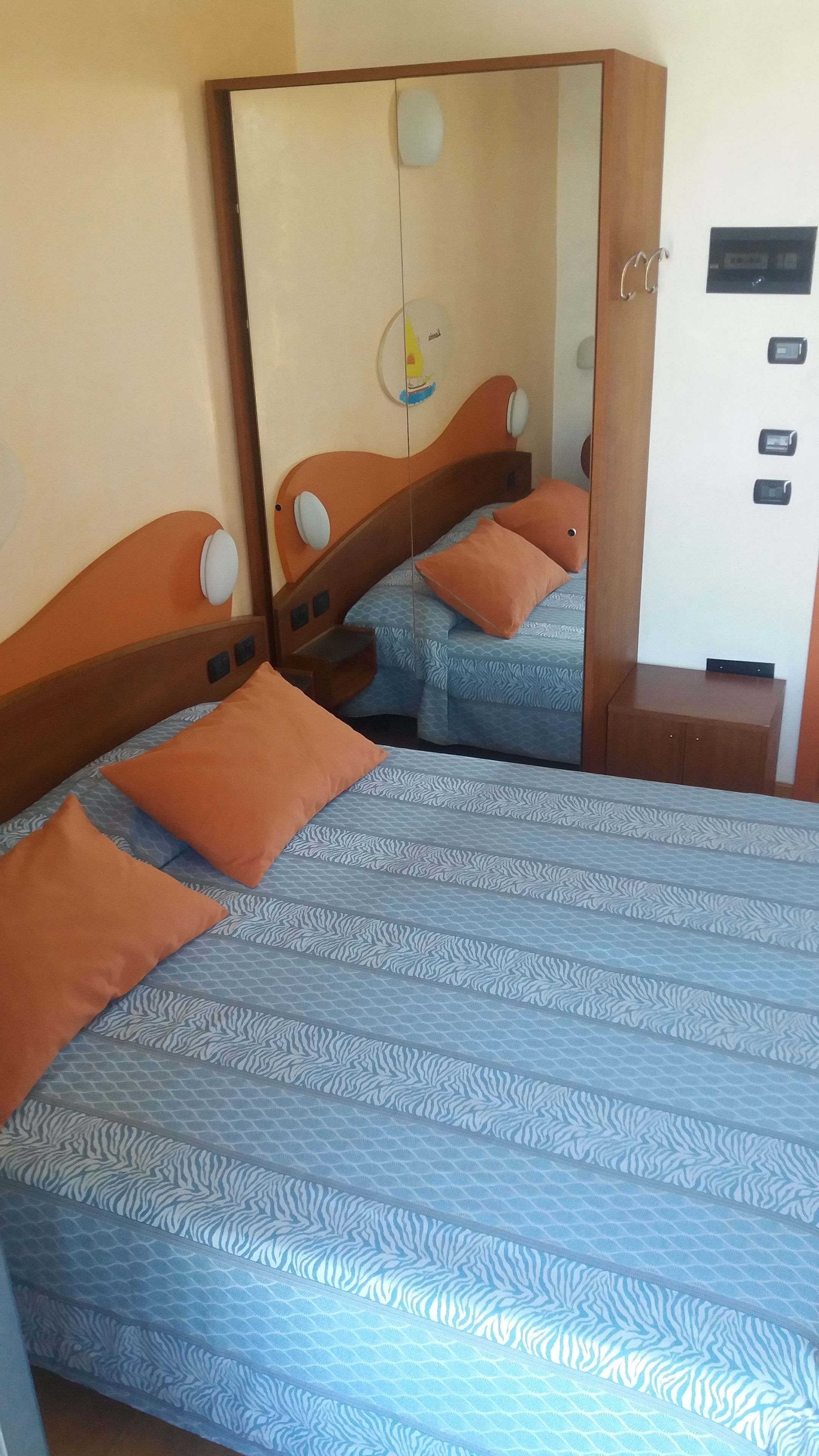 A bedroom with two beds, each with patterned bedding and orange pillows. There is a wardrobe with a mirror and a small desk with a computer.