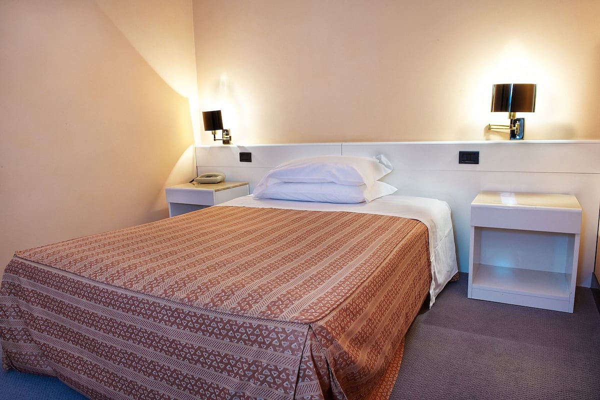 A hotel room with a neatly made bed, two bedside tables with lamps, and a wall-mounted light fixture.
