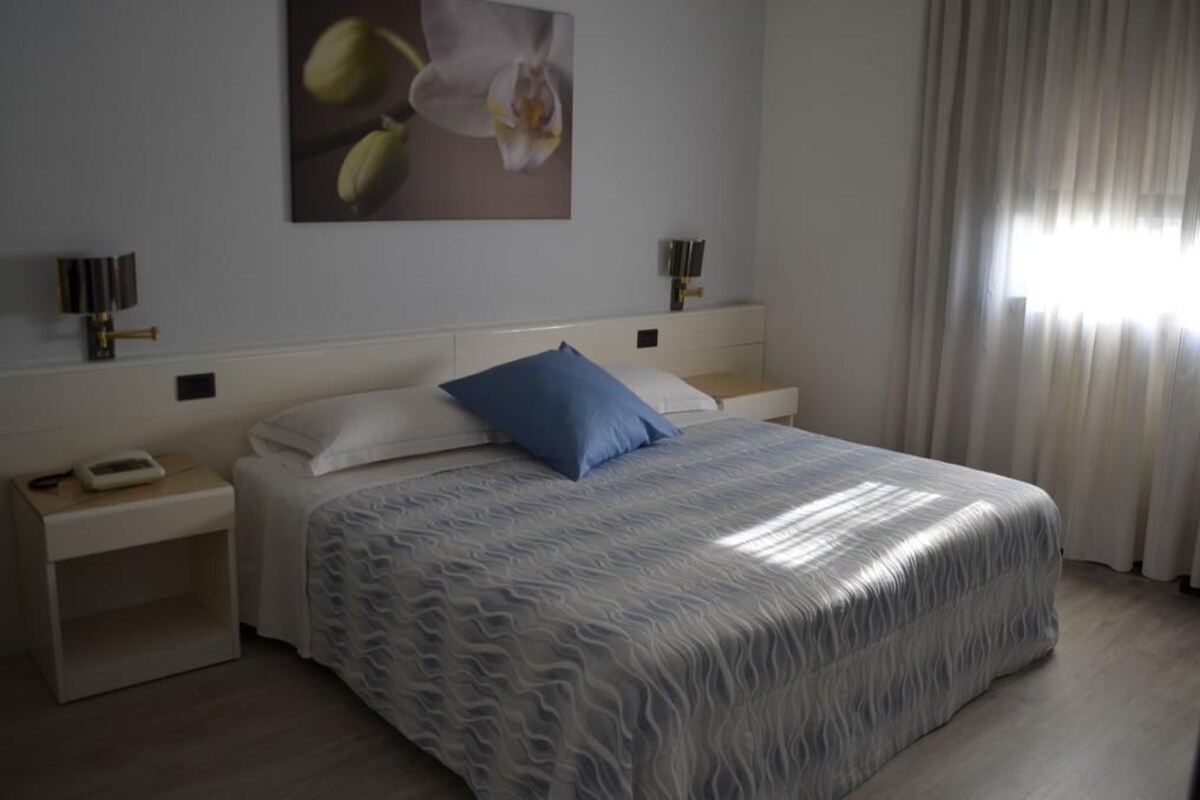 A bedroom with a bed, two bedside tables, a nightstand, a wall-mounted lamp, and a window with curtains.