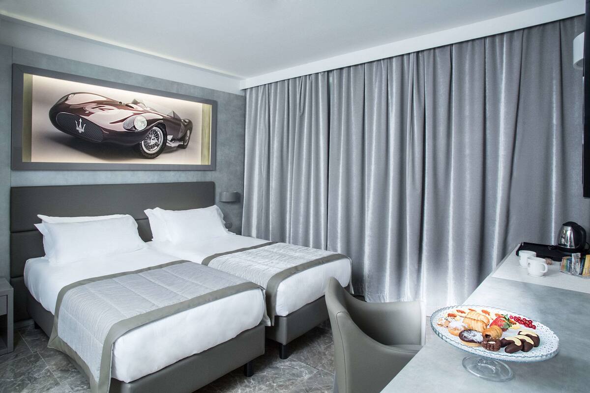 A hotel room with two beds, a dining table with a dessert plate, a coffee machine, and a large window with curtains.