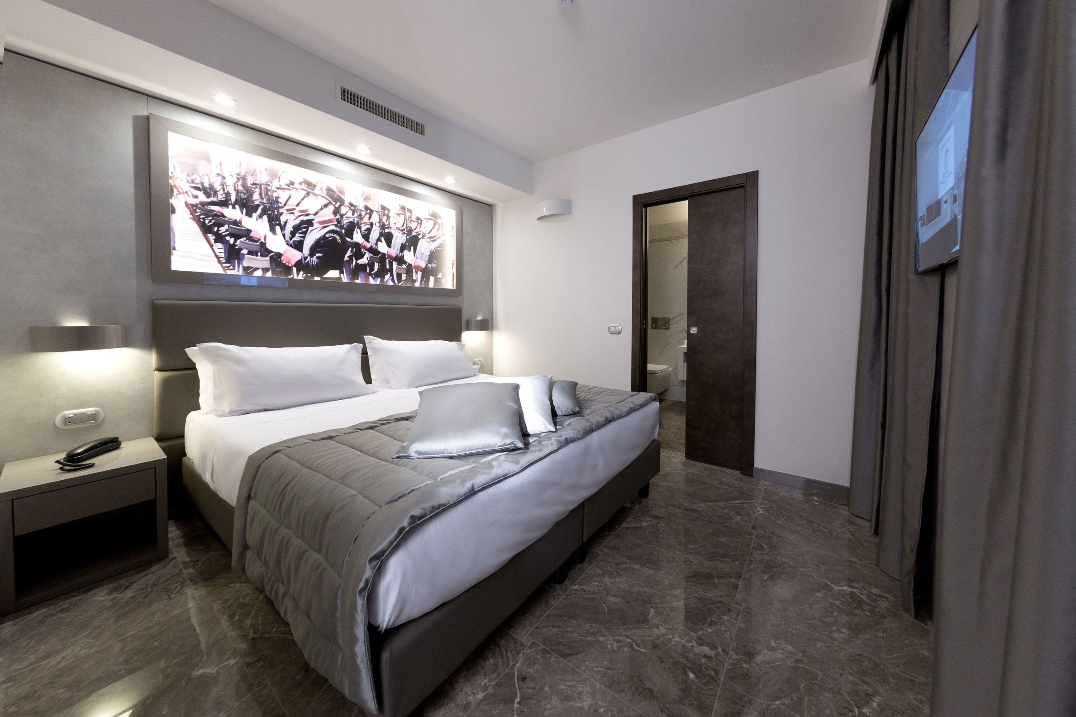 Een moderne slaapkamer met een groot bed, nachtlampen, een aan de muur bevestigde televisie en een deur naar een andere kamer.