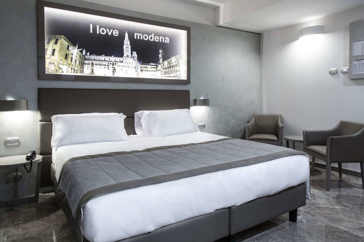 A modern hotel room with a large bed, two bedside tables, a chair, and a wall-mounted TV displaying a cityscape.