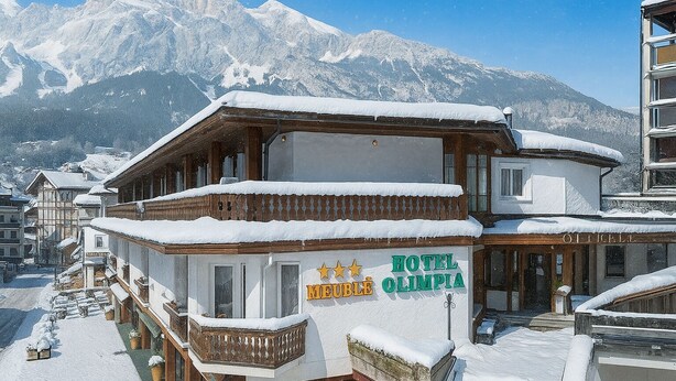 Hotel Olympia with a snow-covered exterior, wooden balconies, and a view of snow-capped mountains in the background. - Hotel Olimpia (Cortina d'Ampezzo)