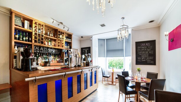 A bar area with a wooden counter, shelves of bottles, and a seating area with tables and chairs. - Victoria Park House Hotel (Edinburgh)