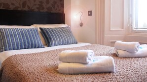 A neatly made bed with striped pillows, a fluffy beige blanket, and rolled towels on a carpeted floor. - Victoria Park House Hotel (Edinburgh)