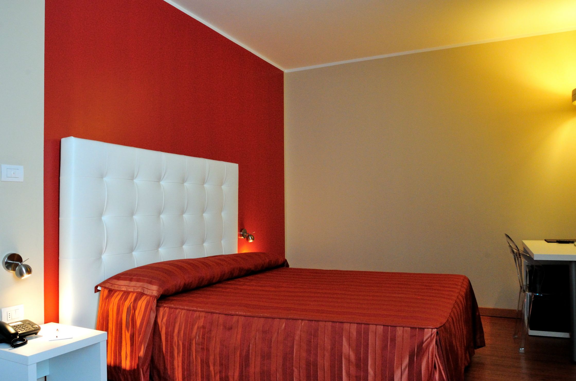 Ein Hotelzimmer mit rotem und weißen Dekor, einem weißen, gepolsterten Kopfteil, einer roten Bettdecke mit Streifen, einem Nachttisch mit Lampe sowie einem Schreibtisch mit Stuhl.