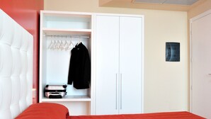 Een slaapkamer met een rood bedovertrek, een wit hoofdeinde, een witte garderobe met kledingroede en een klein zwart object op het onderste plankje.