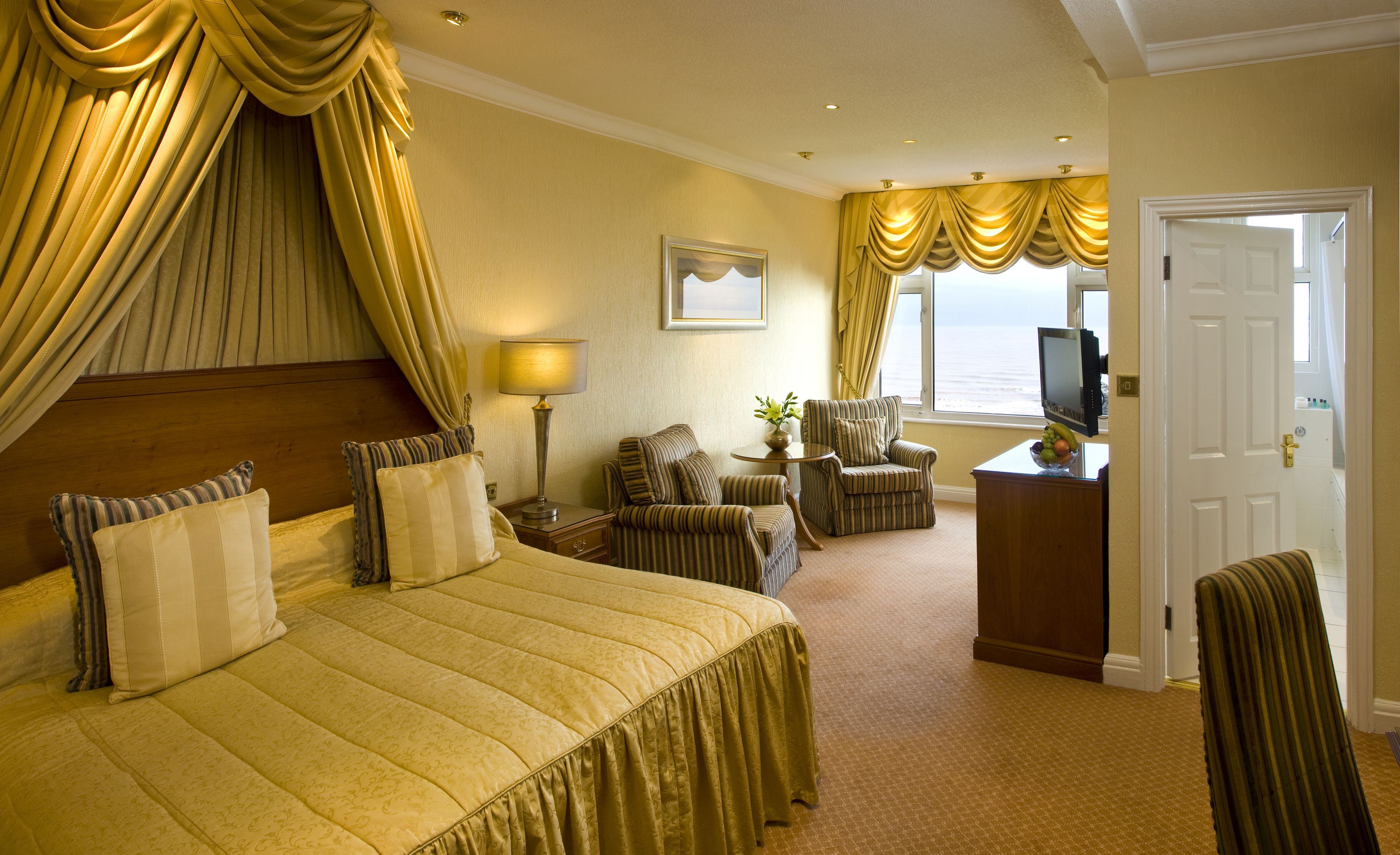 A hotel room with a large bed, a television, a desk with a fruit bowl, and a view of the ocean.