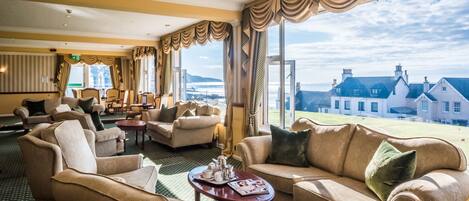 A hotel room with a view of a coastal town, furnished with leather sofas, armchairs, and a coffee table.