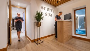 Uma área de recepção com balcão de madeira, um vaso com planta e um letreiro na parede com a inscrição "Source Spa".