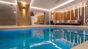 Piscina interior moderna com água azul cristalina, área de descanso com sofá e poltronas, e parede de madeira com ripas verticais.