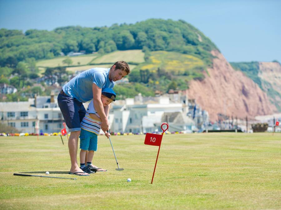 Um campo de golfe com uma bandeira marcada com o número '10' em primeiro plano, uma pessoa vestindo uma camisa e bermuda azuis, e uma criança com roupa listrada.