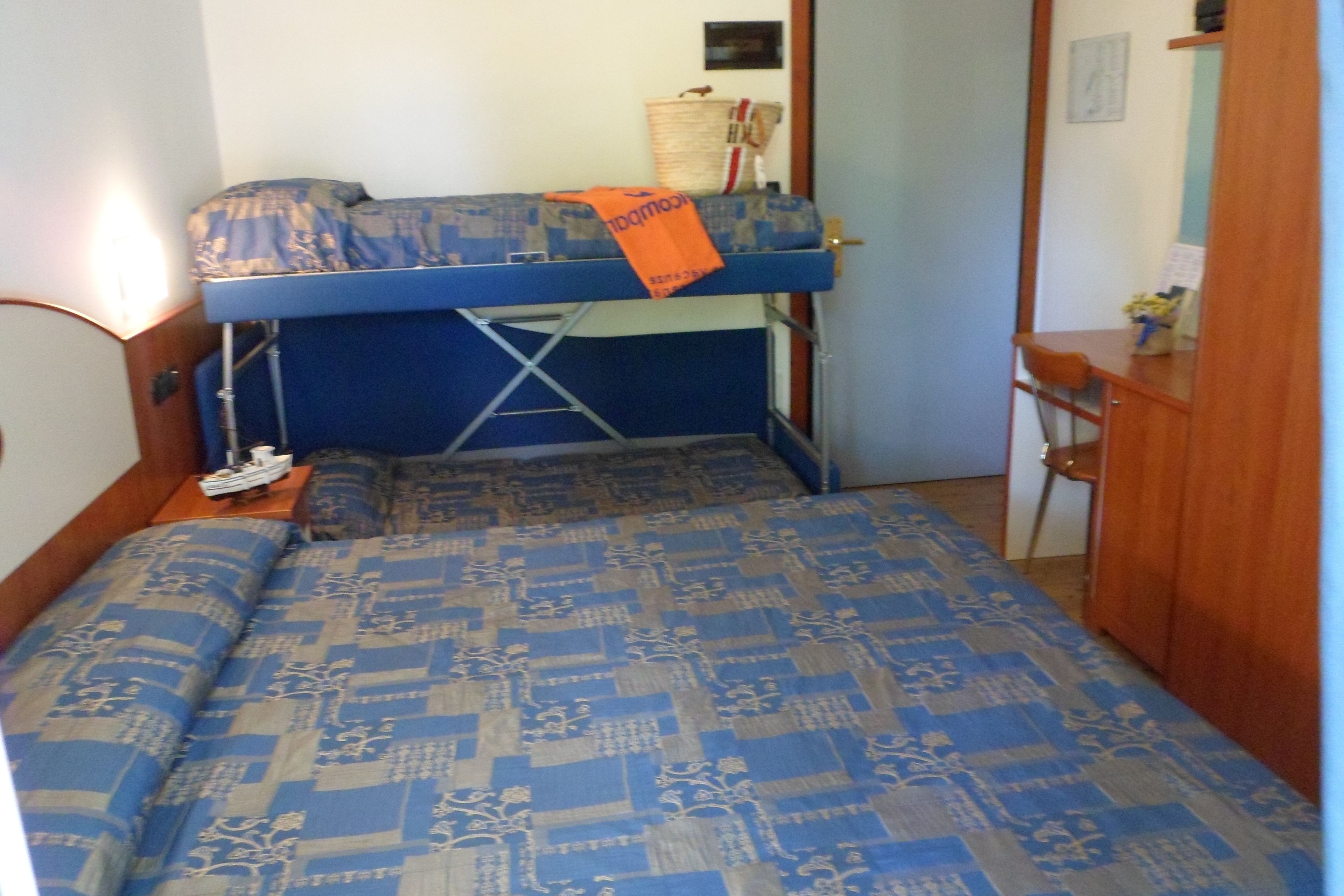 Una cama litera con un colchón azul y beige estampado, una mesita con una cesta y un paño rojo y blanco, y una mesita de noche de madera con una lámpara.