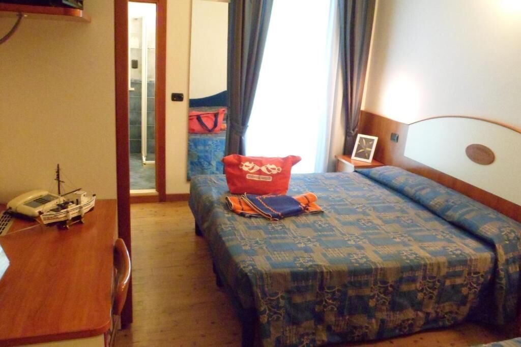 A hotel room with a bed, a desk with a telephone and a ship model, a red pillow, and a framed picture on the bedside table.