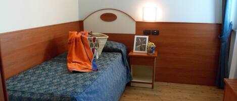 A small, single-bed room with a wooden headboard, a bedside table with a lamp, and a small shelf with items.