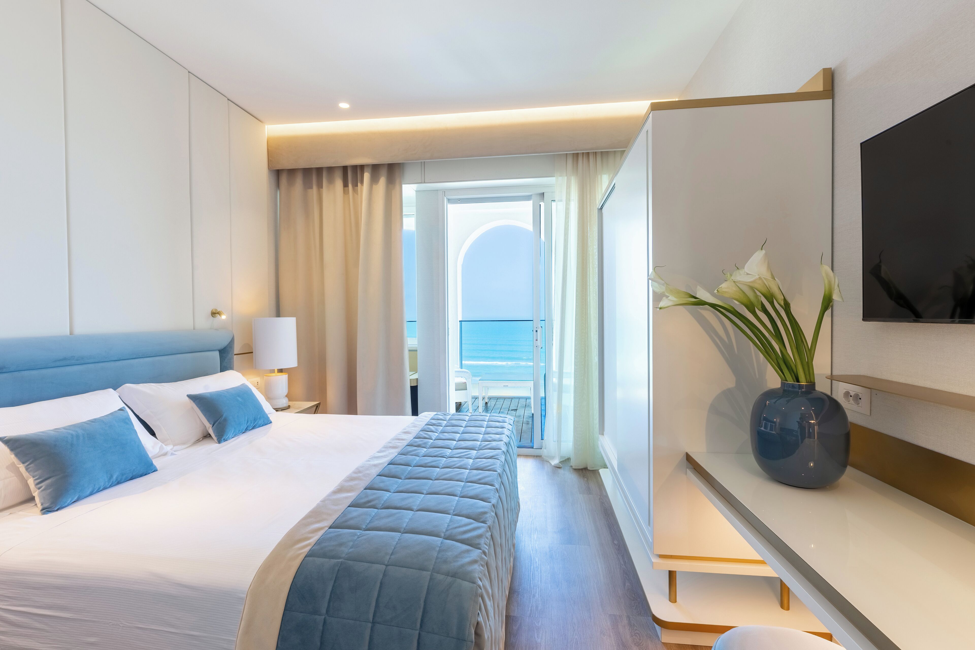 A hotel room with a bed, a desk, a TV, and a view of the sea.