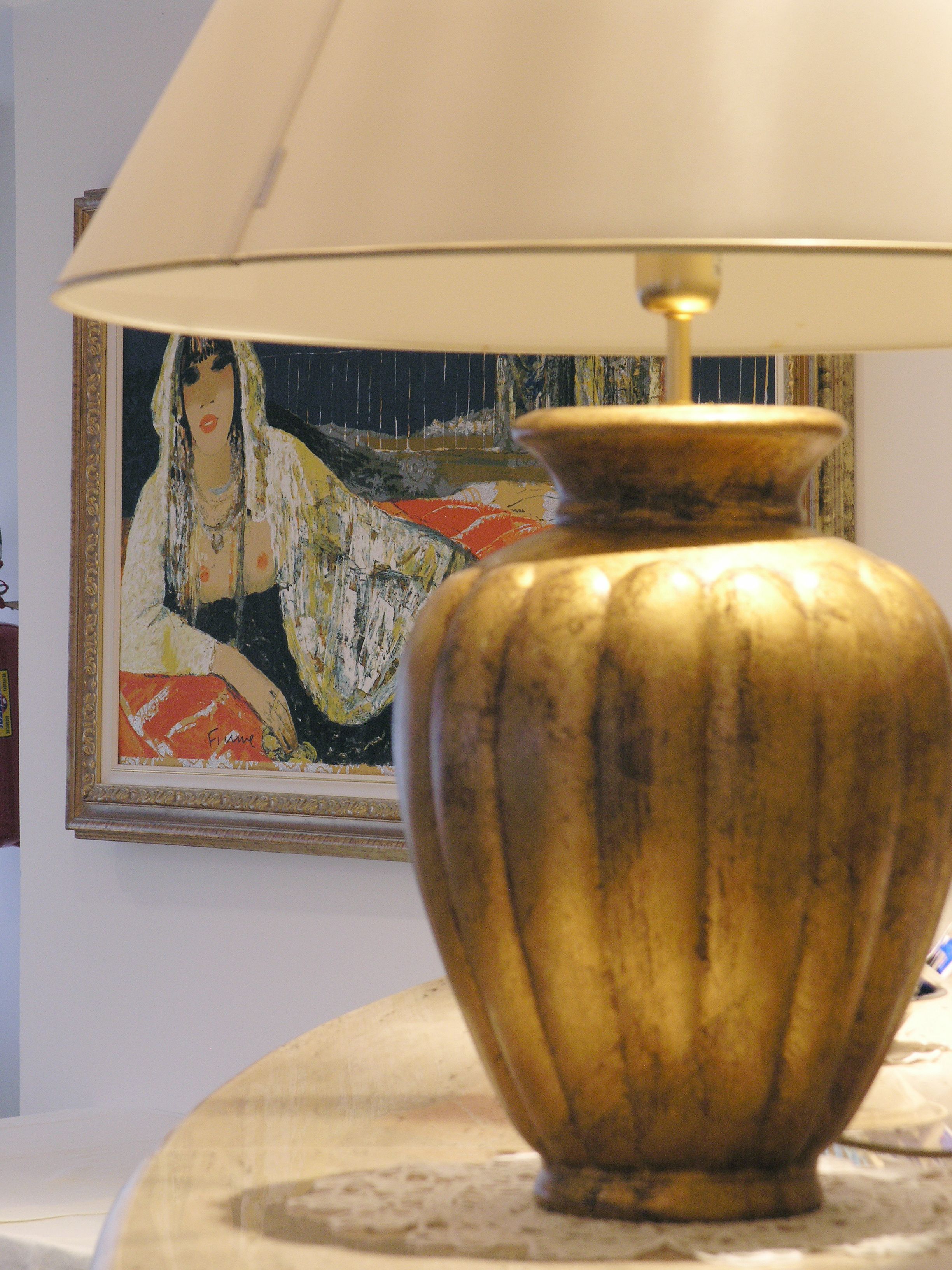 A lamp with a golden base and a white lampshade on a table. In the background, there is a framed painting on the wall.