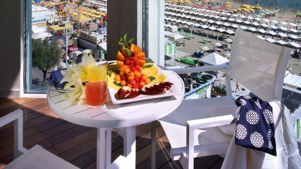 A balcony with a table set for breakfast, overlooking a beach with umbrellas and people. - Tiffany's (Riccione)