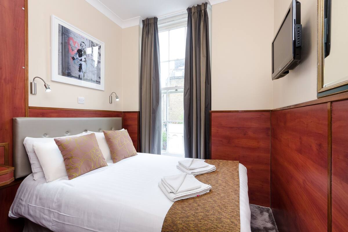 Habitación de hotel con cama, televisor montado en la pared y ventana con cortinas.