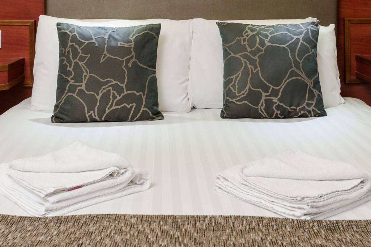 Habitación de hotel con cama, dos almohadas, cuadro con vista urbana enmarcado, dos luces empotradas en la pared y cabecero de madera.