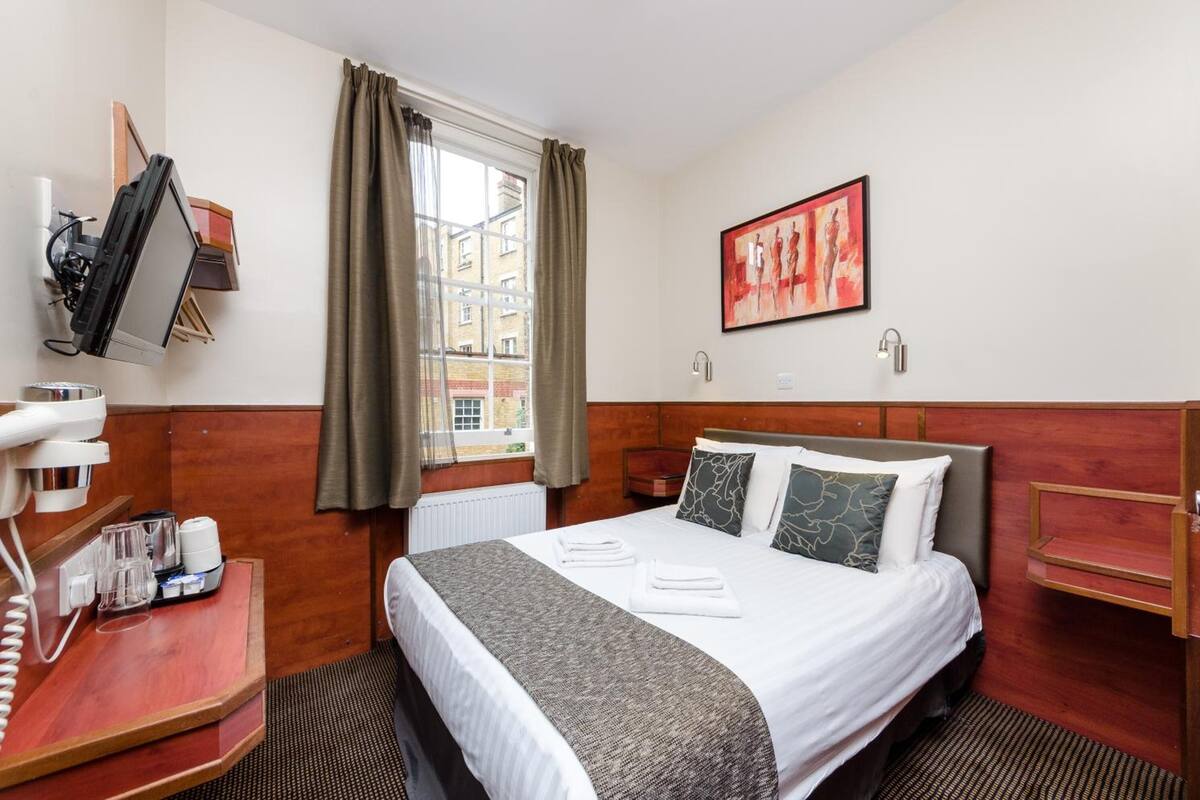 Habitación de hotel con cama, escritorio con teléfono y gafas, ventana con cortinas y un cuadro en la pared.