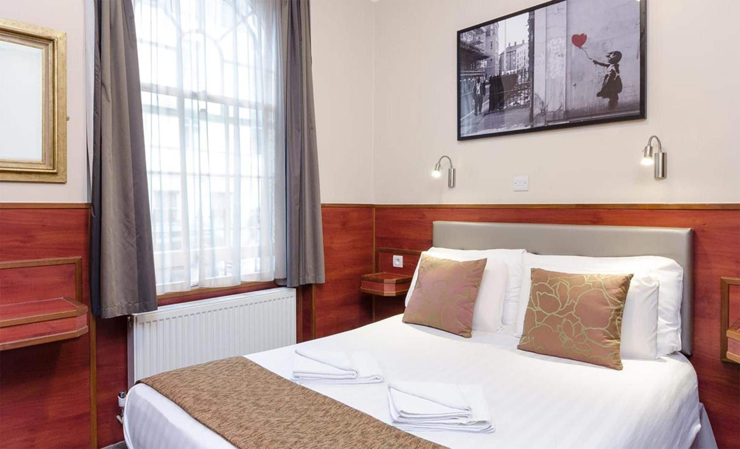 A hotel room with a large bed, a window with curtains, a painting on the wall, and a nightstand.