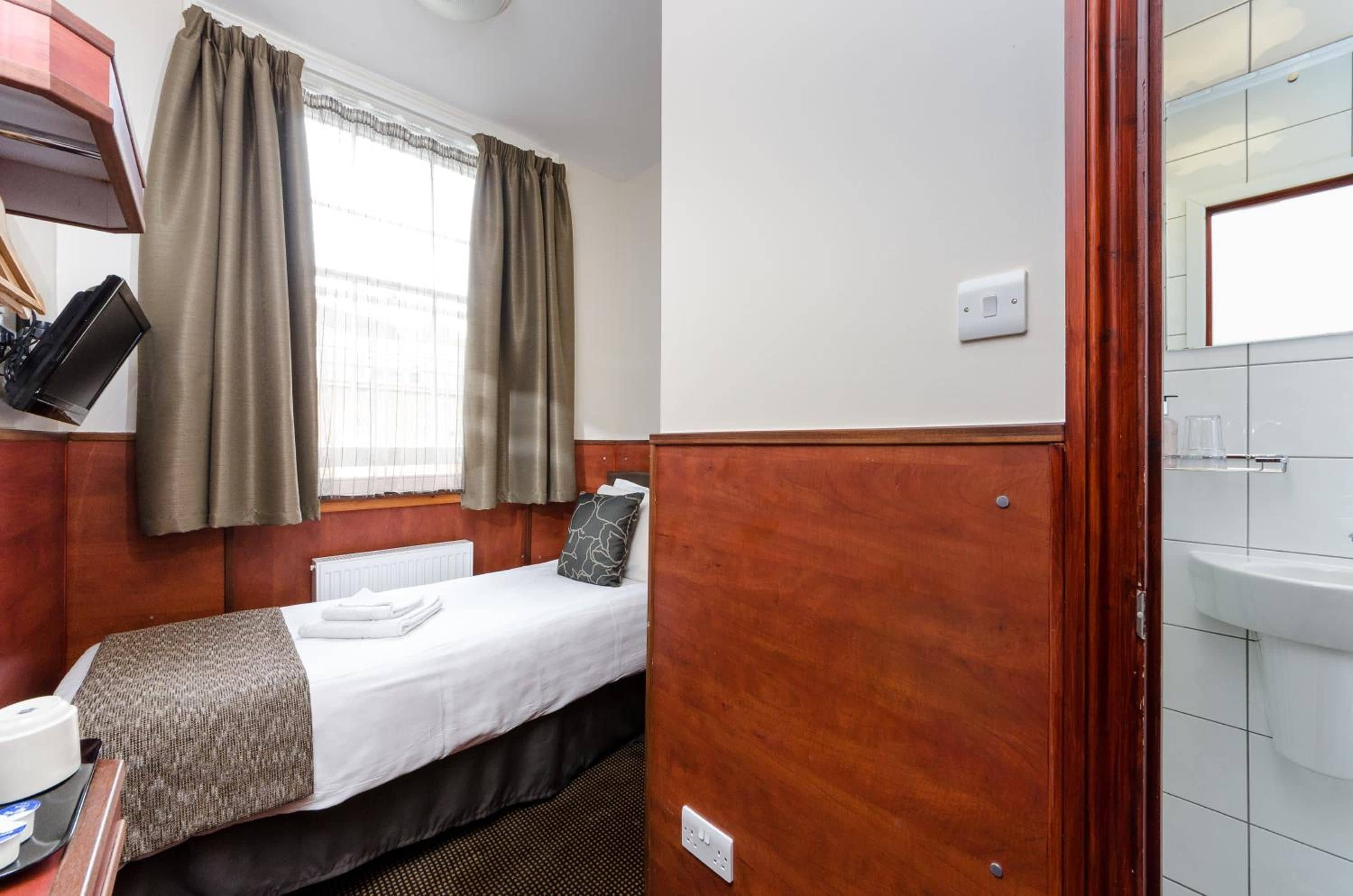 Habitación de hotel pequeña con una cama individual, una mesita de noche, un enchufe, una ventana con cortinas y vistas al exterior.