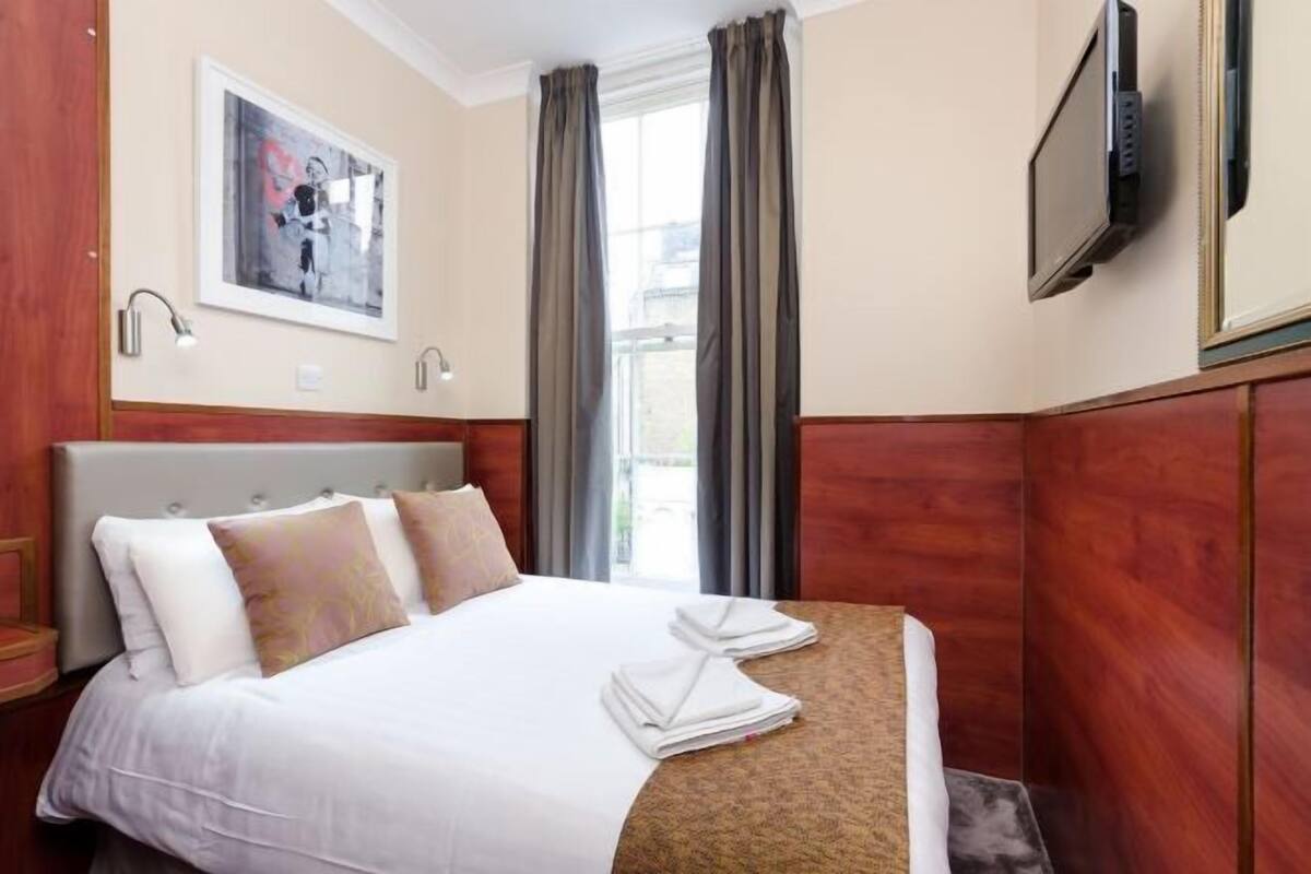 A hotel room with a bed, a flat-screen TV, a framed picture on the wall, and a window with curtains.