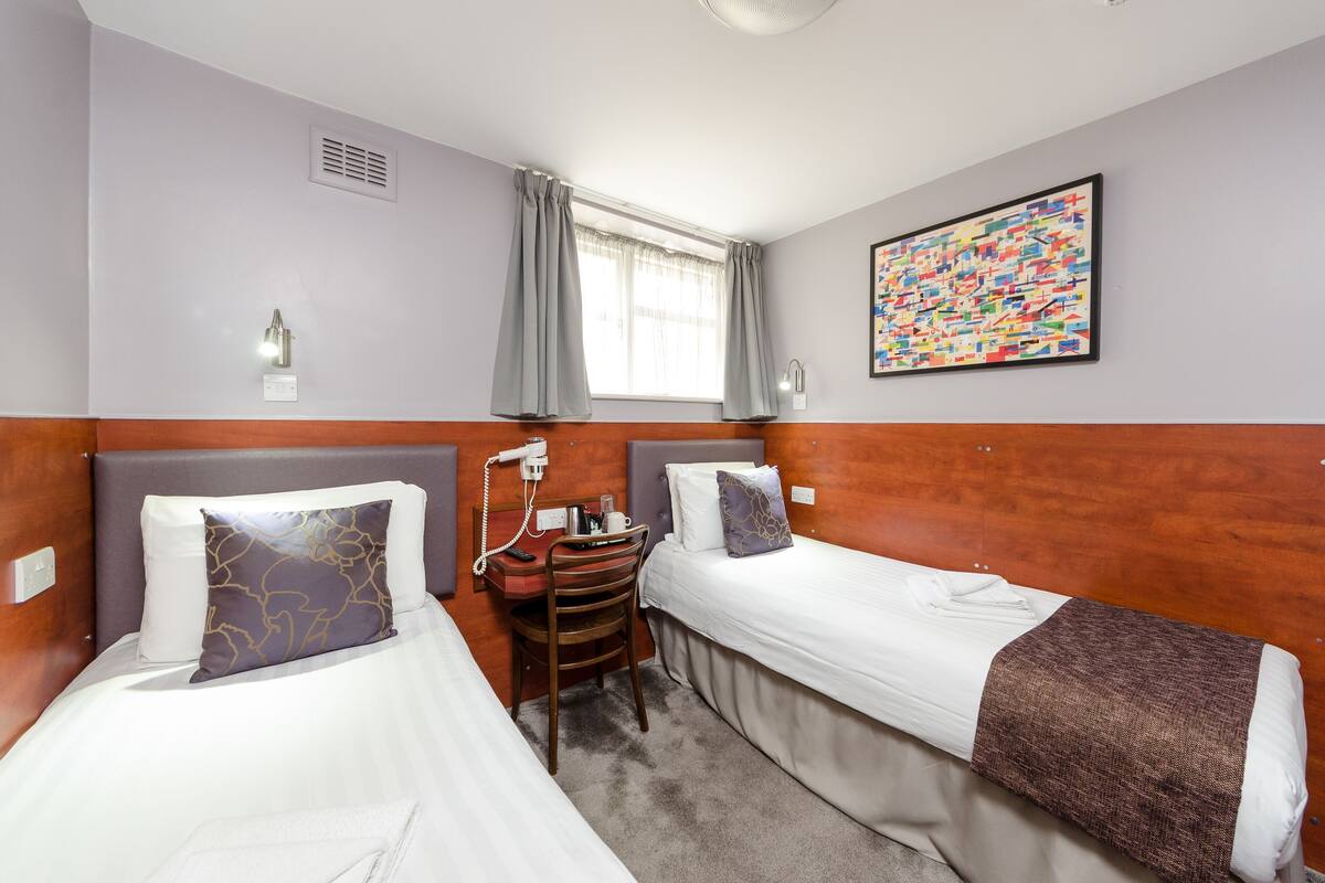 Habitación de hotel con dos camas individuales, un escritorio con silla, una ventana con cortinas, una lámpara fijada a la pared y un cuadro abstracto de colores.
