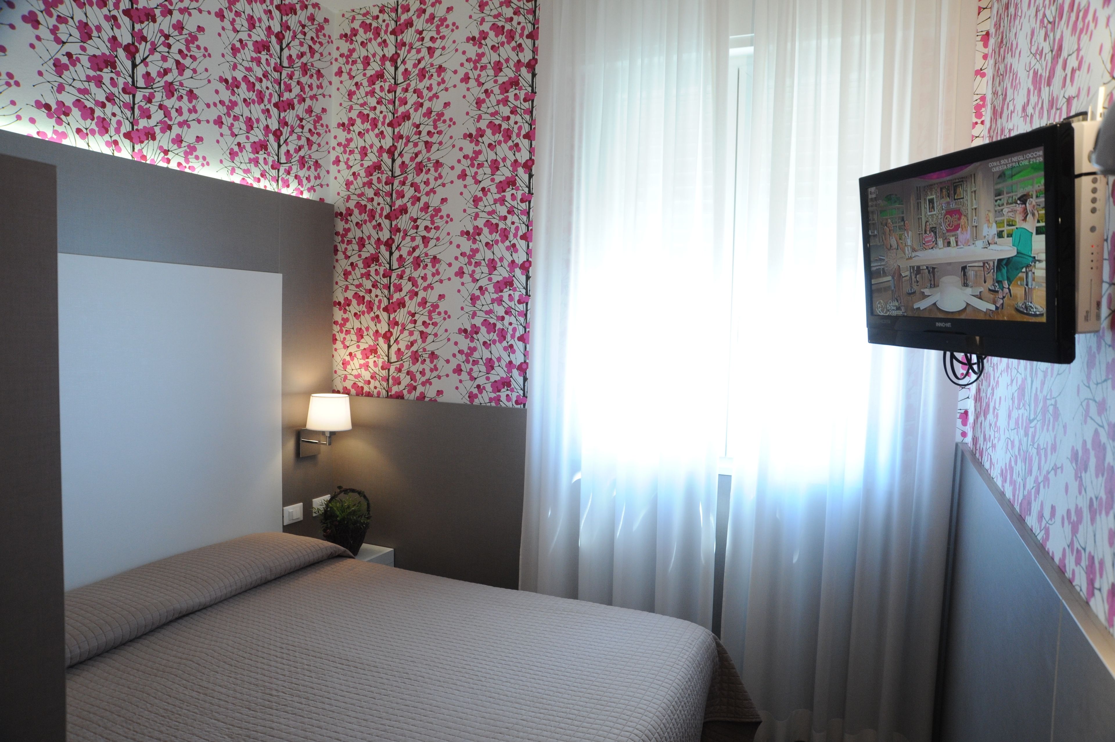 A hotel room with a bed, a wall-mounted TV, a bedside lamp, and a window with sheer curtains.