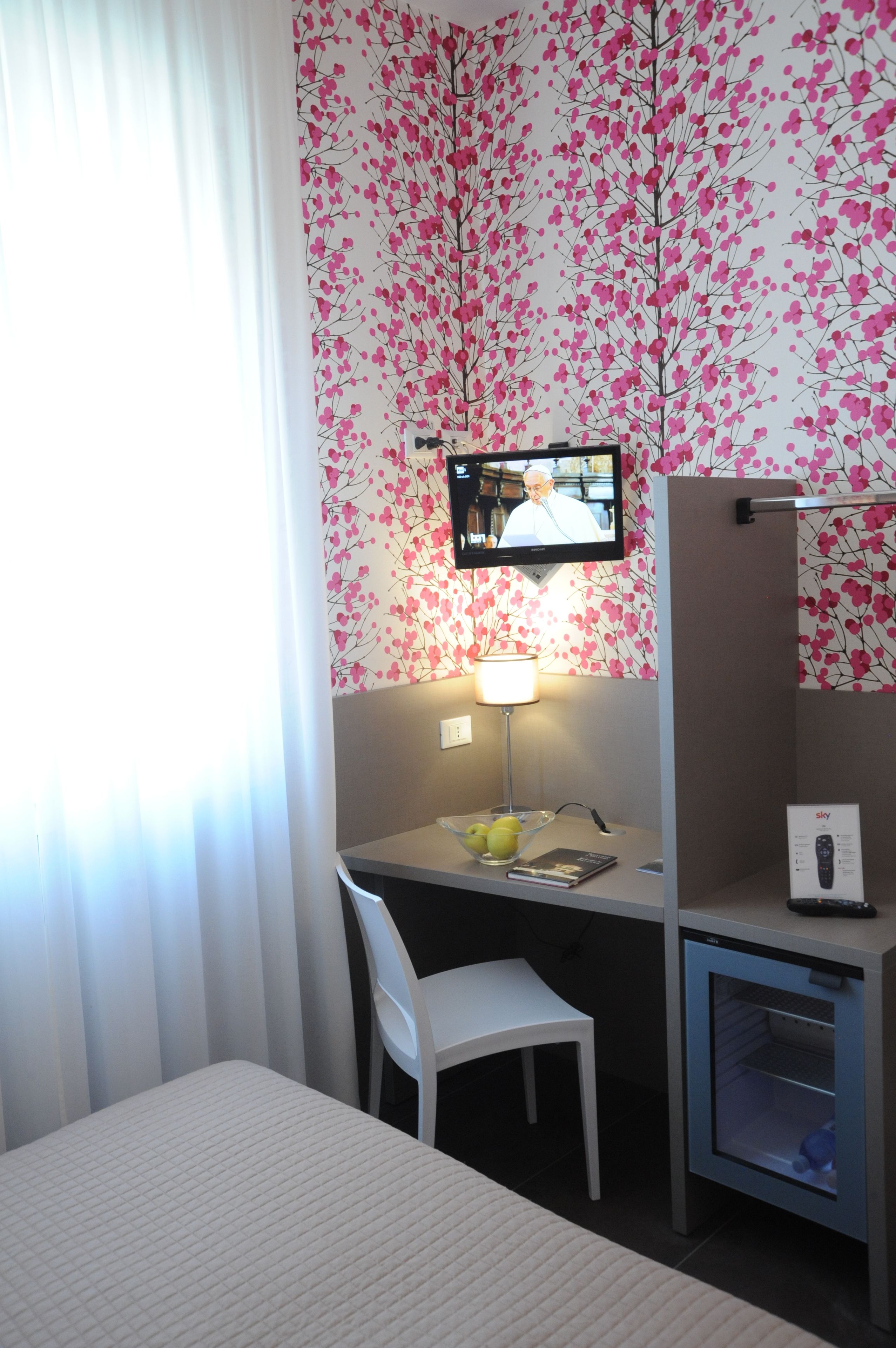A hotel room with a bed, a desk with a chair, a television mounted on the wall, a small refrigerator, and a window with curtains.