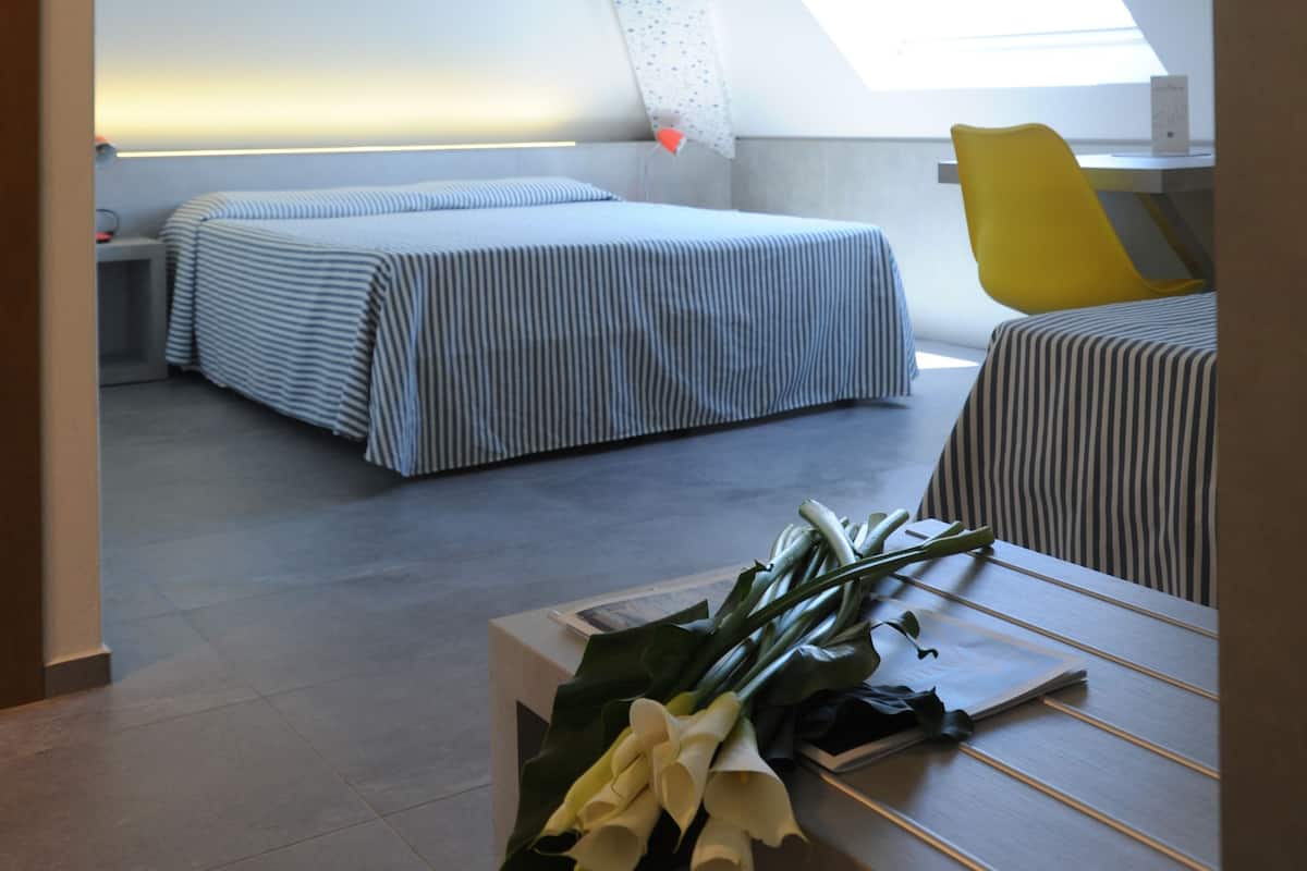 Ein modernes, kompaktes Zimmer mit schrägem Dach, zwei Betten mit gestreifter Bettwäsche, einem kleinen Tisch mit Blumenstrauß und einem hellen Dachfenster.