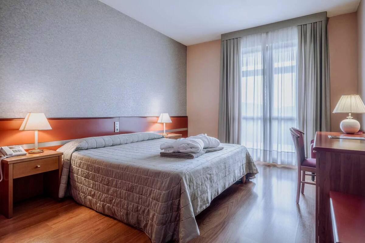 Une chambre d’hôtel avec un grand lit, deux lampes de chevet, un bureau avec une chaise et une grande fenêtre avec des rideaux.