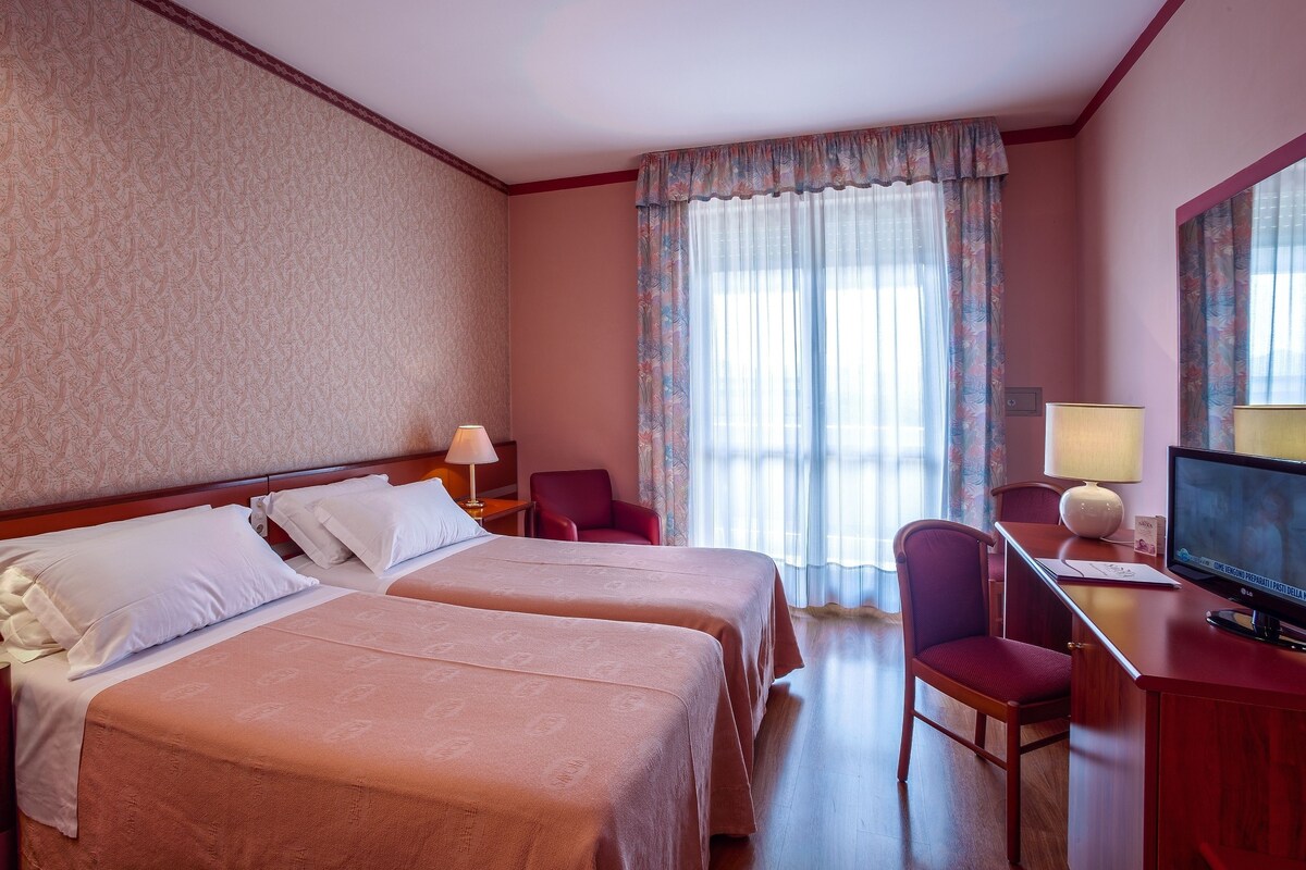 Een hotelkamer met twee bedden, een bureau met een televisie, een stoel en een raam met gordijnen.