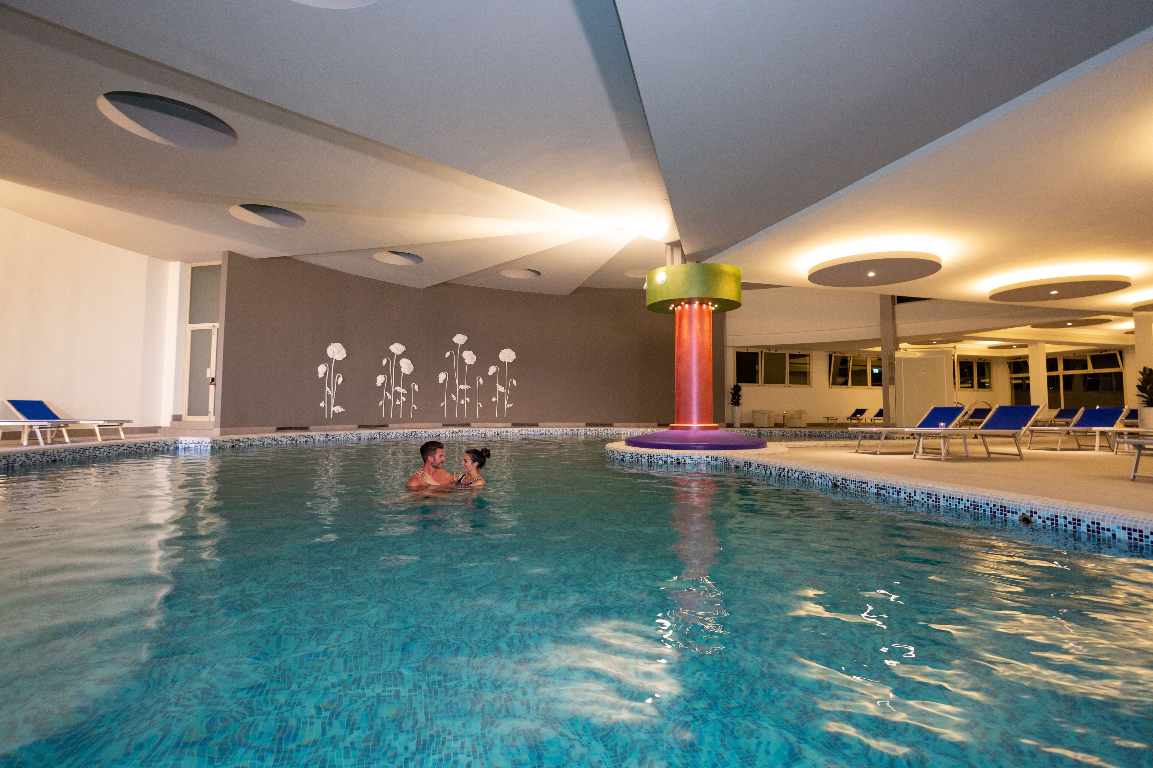An indoor swimming pool with blue water, a decorative wall with white floral patterns, and several blue lounge chairs around the pool.