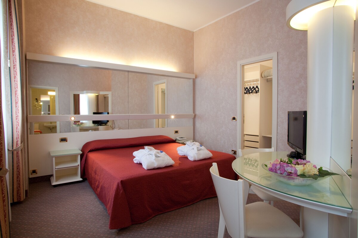 Une chambre d’hôtel comprenant un lit, un bureau avec un plateau en verre, une chaise, un téléviseur et un miroir.